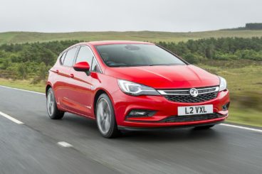 bright red vauxhall astra driving on tarmac road with fields in the background