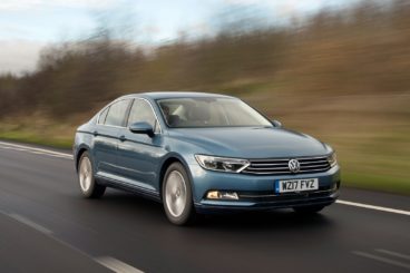 metallic grey volkswagen passat saloon driving on tarmac road with green trees in the background