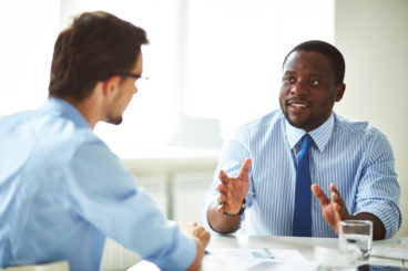 two businessmen wearing blue shirts and ties sitting talking over a table