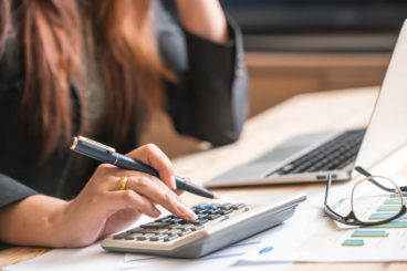 woman with brown hair working out calculations next to a laptop with a pen in her hand