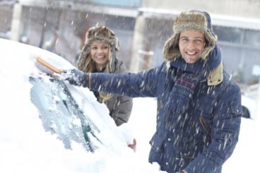 couple in coats and winter hats int he snow smiling while clearing snow off their car