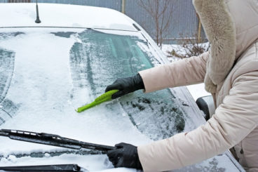 person in cream coat wearing gloves scraping snow off their windscreen