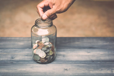 glass money jar on wooden table with hand putting more money in