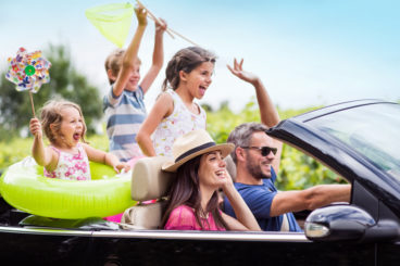 two adults and three young children in a convertible travelling on holiday with rubber rings and windmills