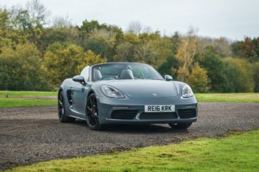 grey porsche boxster parked on gravel track with green fields on either side and trees in the background