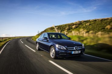 dark blue Mercedes Benz C-Class AMG Saloon driving down tarmac road next to grass hills