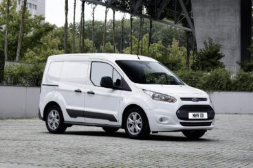 white ford transit connect parked on pebble road with trees and a modern building in the background