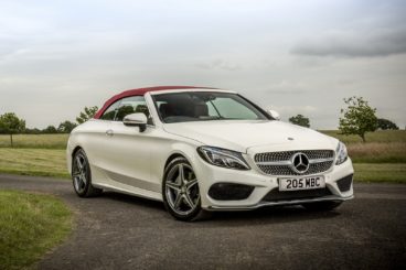 a white mercedes c class cabriolet parked on gravel with green trees and fields in the background