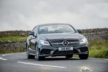 a dark mercedes CLS driving on tarmac road with fields on either side