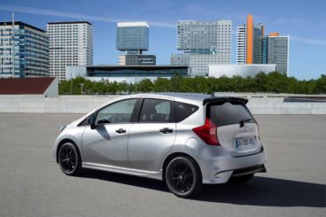 view of the back of a metallic nissan note hatchback parked on tarmac in front of city setting