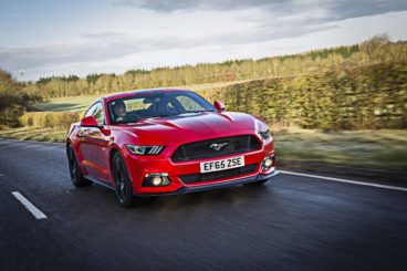 bright red ford mustang fastback driving down tarmac road with fields to the side of it