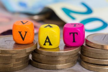 colourful orange, yellow & pink dice spelling out VAT on top of coins with euro notes blurred in the background