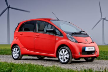 bright red citroen czero parked on gravel with green grass on either side and wind mills in the background