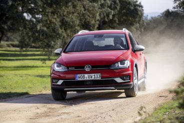 bright red volkswagen golf all track driving down dusty road with green grass and trees in the background