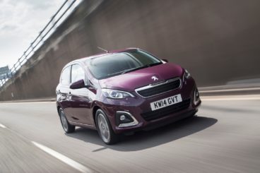 purple coloured peugeot 108 hatchback driving on tarmac road with concrete wall in the background