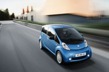 a metallic blue peugeot ion hatchback driving on tarmac road with blurred background