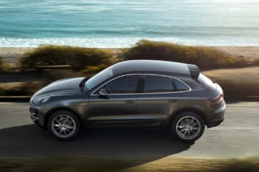 dark mettalic porsche macan estate driving on tarmac road with the beach in the background