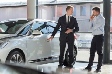 man in a suit showing a man in a shirt and trousers a silver car