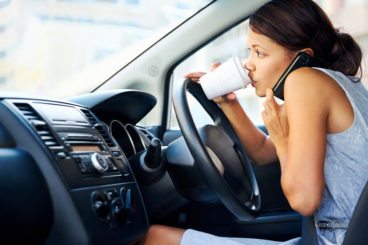 woman driving in a grey dress while drinking coffee and on the phone