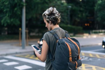 person in a green t shirt with short hair and a backpack listening to music and on their phone while at a crossing