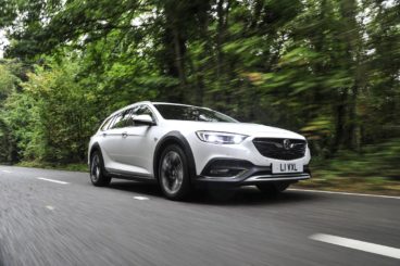 a metallic white Vauxhall Insignia Country Tourer driving down tarmac road with green forest trees in the background