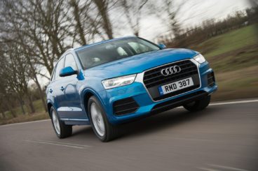 a metallic blue audi q3 diesel estate driving on tarmac road with winter trees in the background