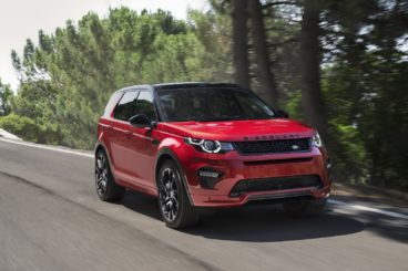 a metallic red land rover discovery sport driving along tarmac road with blurred trees in the background