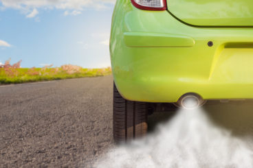 a metallic green car driving up tarmac road with grey smoke coming out of the exhaust pipe
