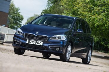 a dark metallic coloured bmw 2 series gran tourer parked on gravel with green tree in the background