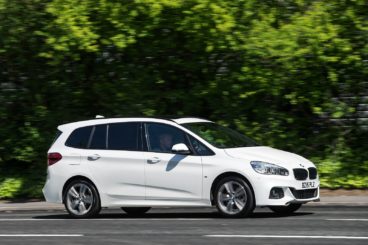 a sold white bmw 2 series diesel gran tourer driving on tarmac road with green trees in the background