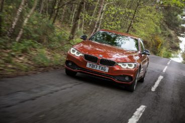 a metallic bmw 4 series diesel coupe driving on tarmac road with green trees in the background