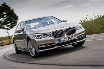 a metallic bmw 7 series diesel saloon driving on tarmac road with green grass and trees in the background