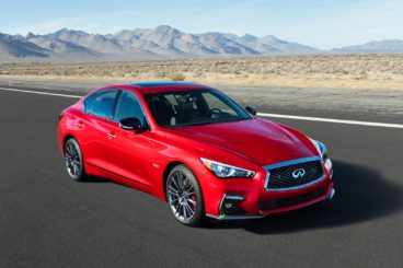 a bright red infiniti Q50 saloon driving on tarmac road with dessert setting in the background