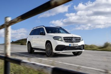 a bright white mercedes GLS class estate driving on tarmac road with fence in the for front