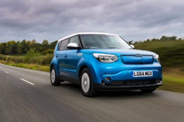 a metallic blue kia soul EV driving on tarmac road with green bushes in the background
