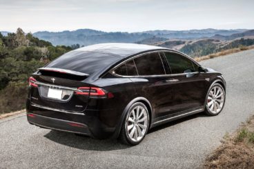 a dark coloured tesla model x parked on gravel with green hills in the background