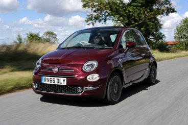 a metallic red fiat 500 driving on tarmac road with green trees in the background