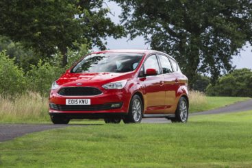 a metallic red ford c max parked on tarmac with bright green grass and trees in the background