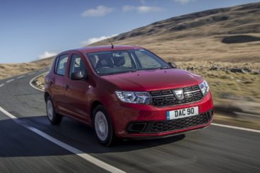 a dark red dacia sandero hatchback driving on tarmac road with grassy hill in the background