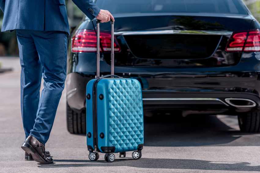A man in a blue suit with brown shoes is holding a light blue suitcase walking towards a black premium car.