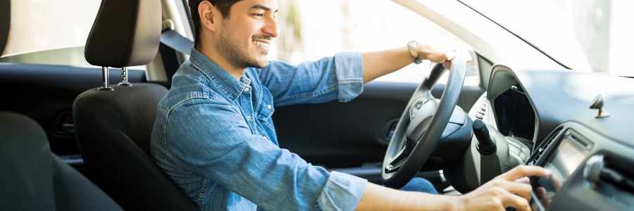 A man wearing a blue denim shirt is in a car with black interior, his hands are on the wheel and he's interacting with the infotainment system