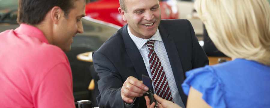 Couple featuring brown haired man and blonde haired lady at a dealership opposite a salesman handing them car keys