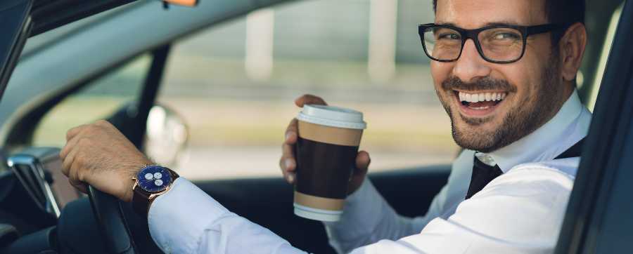 Man wearing glasses and suit sitting in the driver's seat of a car holding a car and smiling