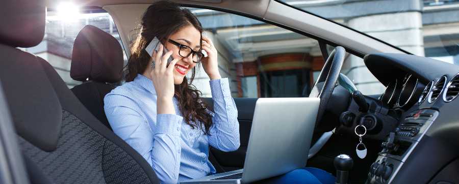 Woman wearing glasses and smart shirt in the driver's seat of a car on the phone smiling whilst looking at her laptop on her lap