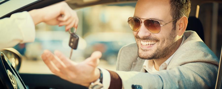Man in a suit smiling in a car with his hand out the window grabbing a key from another hand