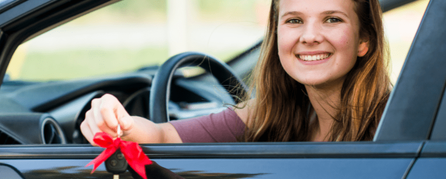 Female smiling sat in the driver's seat of a car holding car keys with a red ribbon attached to it out of the car window - car subscription UK