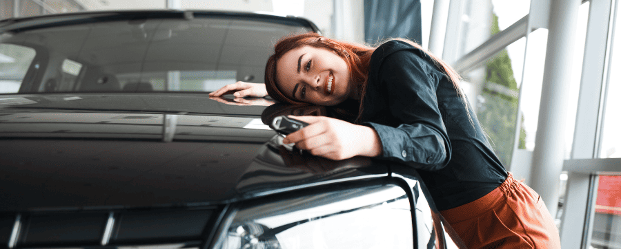 Woman smiling holding car keys whilst leant over hugging a car 